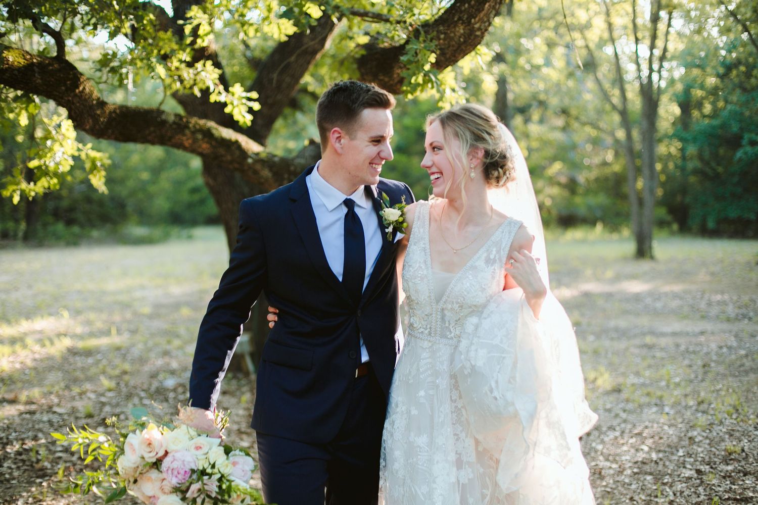 Newlywed couple walks outdoors sharing a joyful moment beneath a tree in a sunlit garden setting.