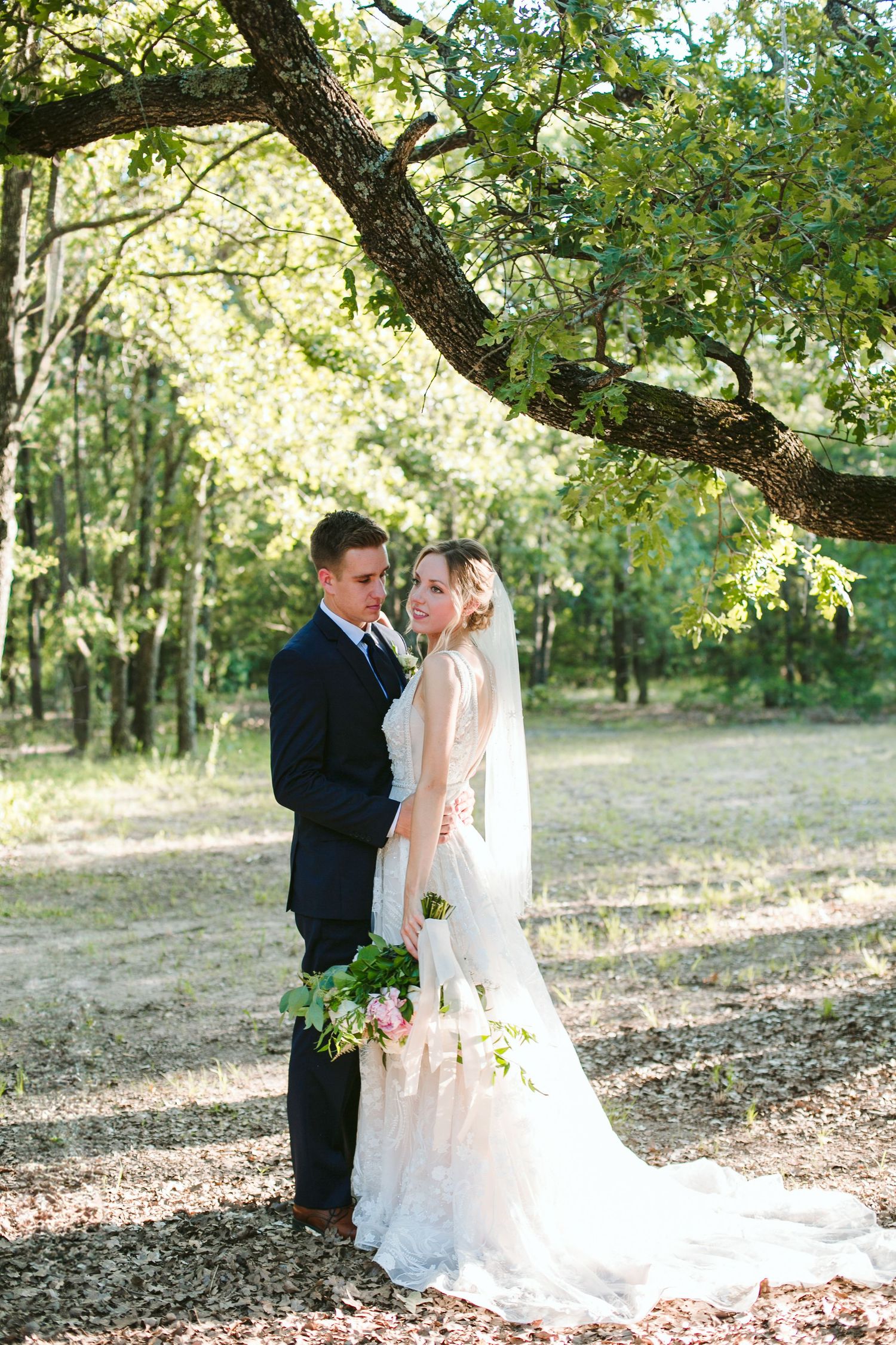 A wedding couple poses under a large tree branch in a sun-dappled forest setting.