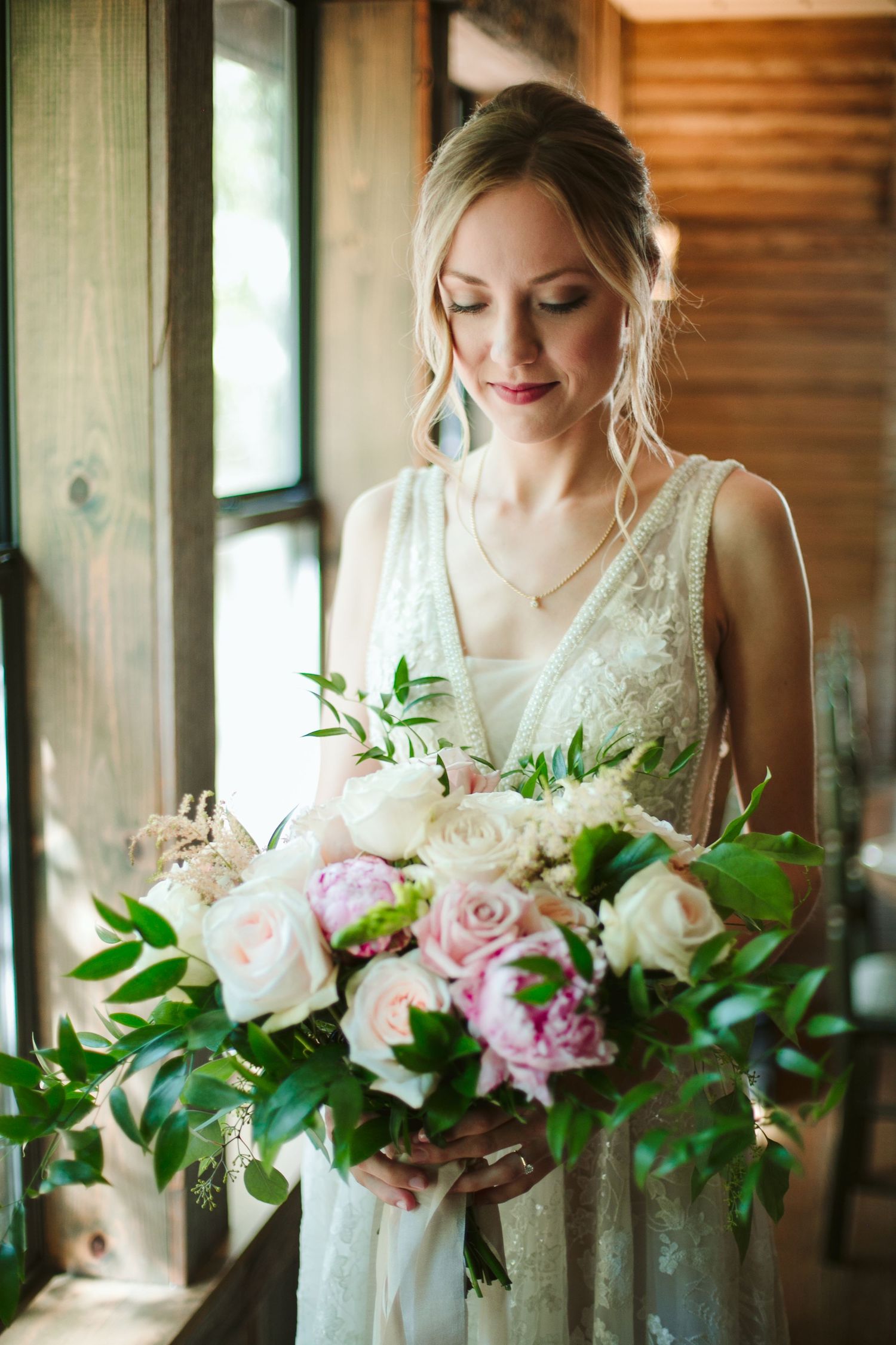 Bride holding romantic bouquet of pink and white roses with greenery by window.
