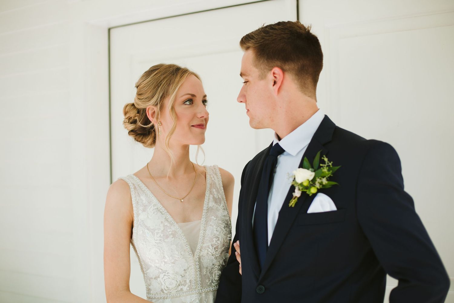 Close-up portrait of bride and groom in formal attire against white interior wall.
