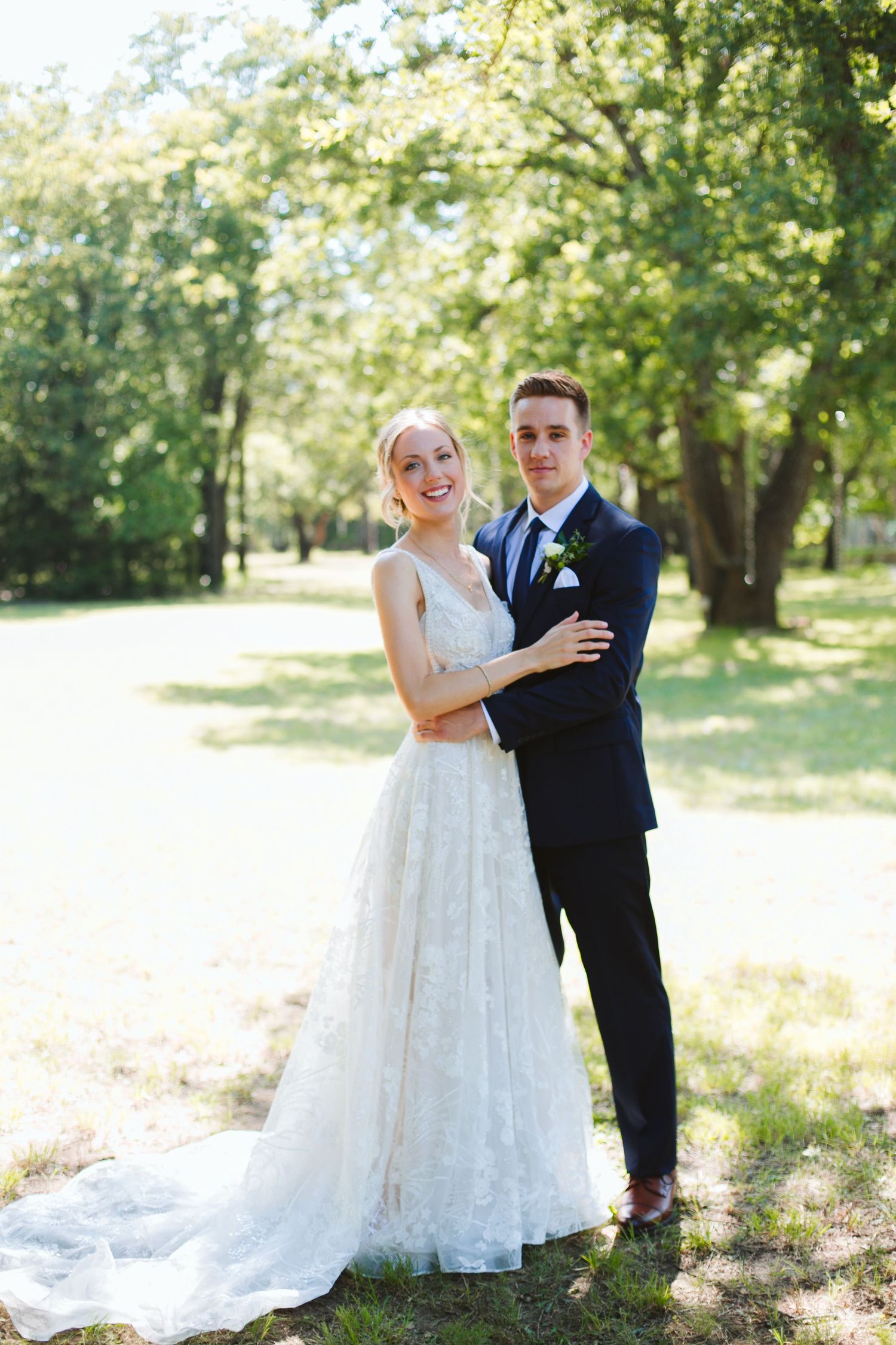 Elegant wedding couple poses in sunlit garden with trees in background.
