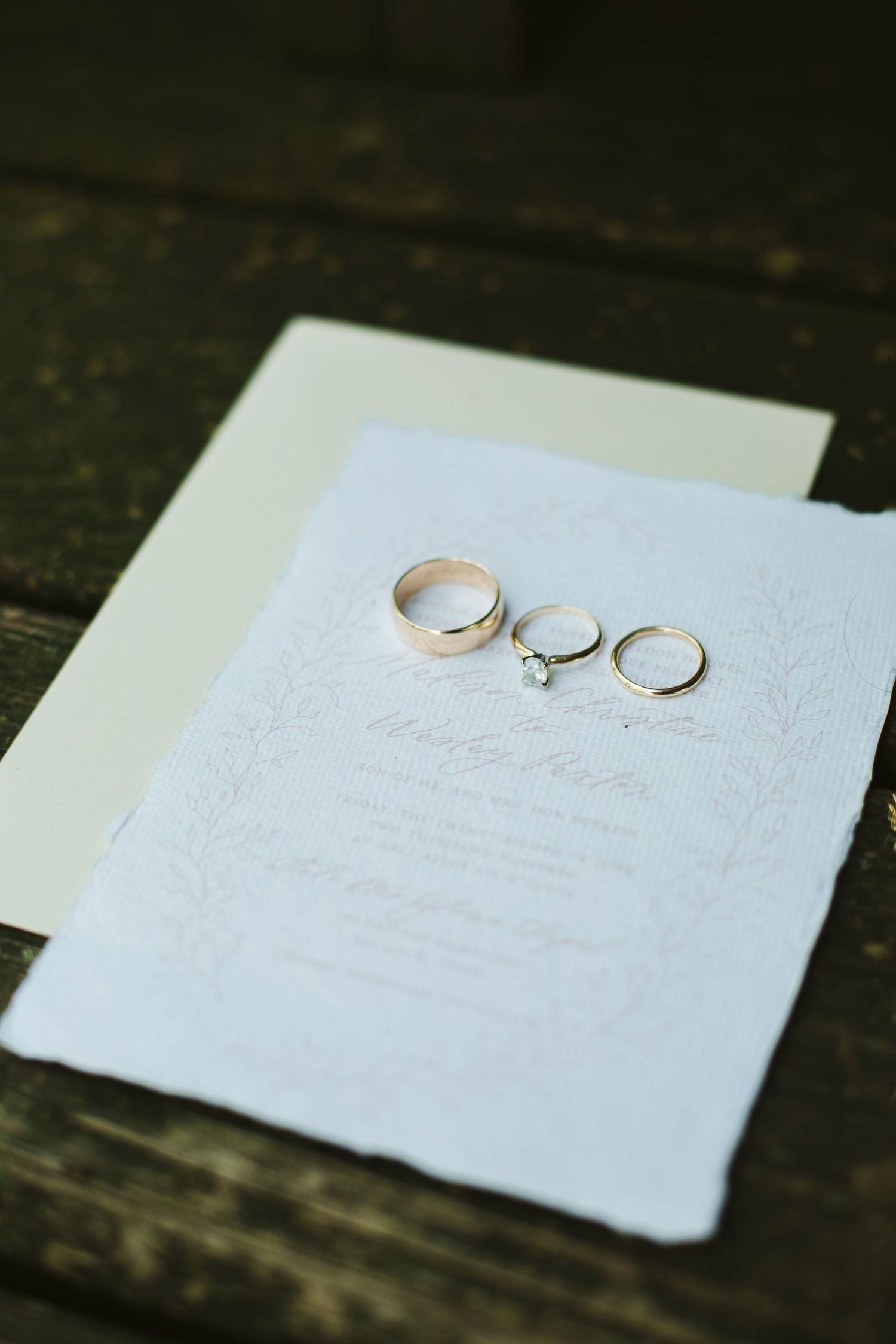 Three wedding rings displayed on white invitation paper against dark wooden surface.