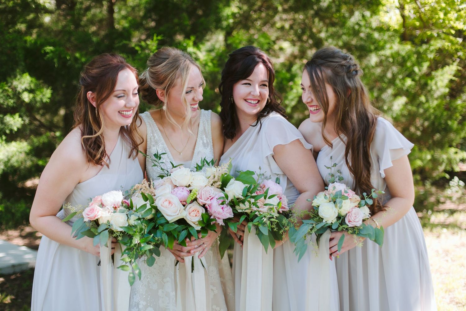 Four bridesmaids in light colored dresses laugh together while holding white and pink floral bouquets outdoors.