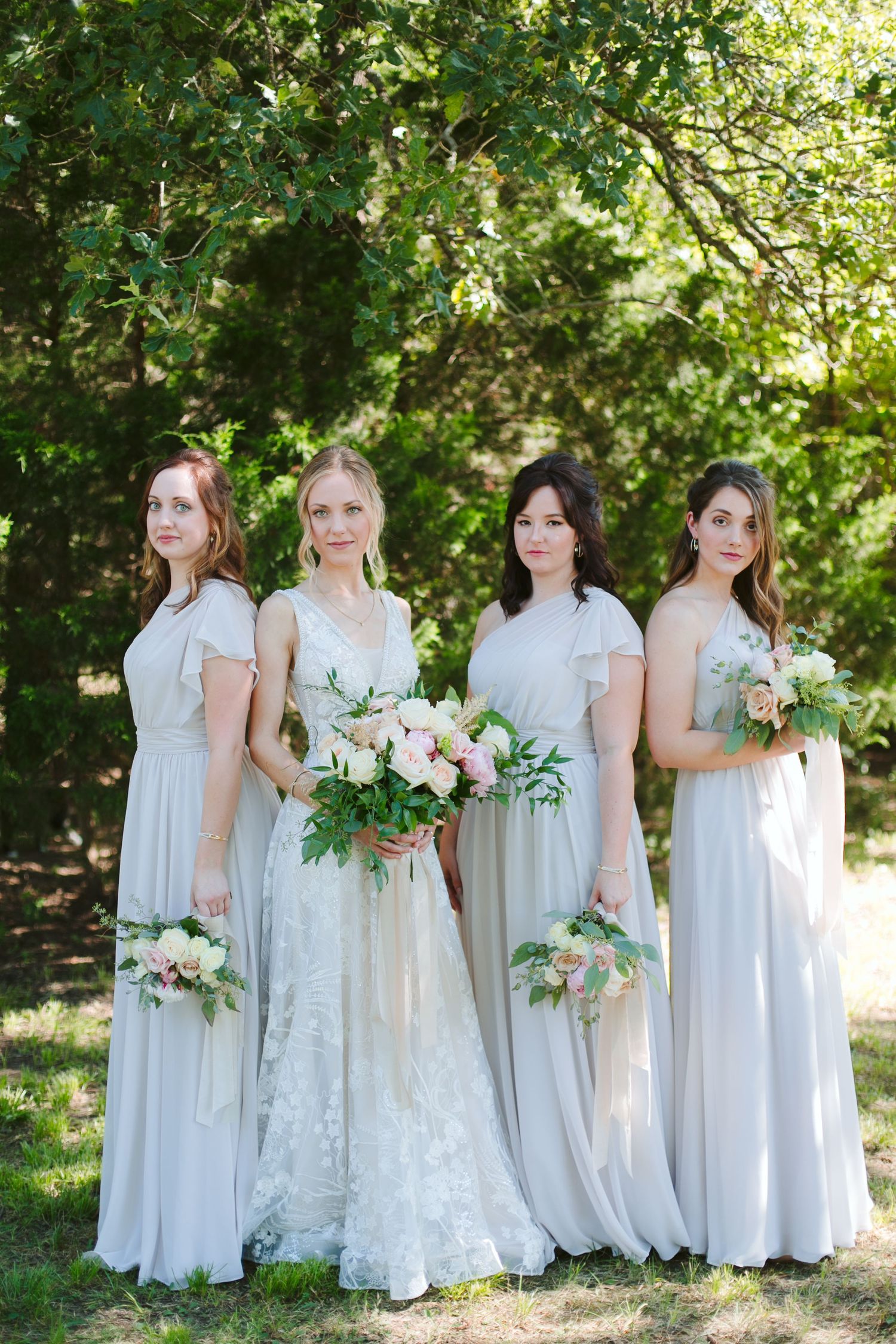 Four bridesmaids in light gray dresses hold bouquets in outdoor garden setting.