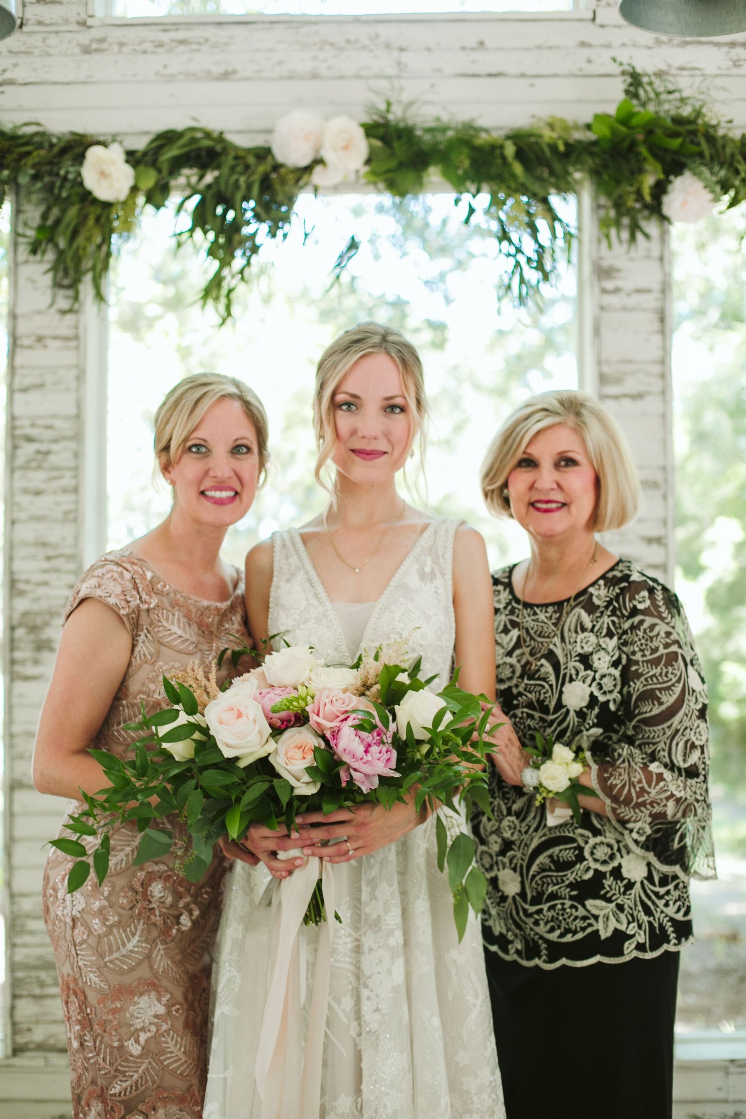 Three elegantly dressed women stand together under a floral archway with roses and greenery at a wedding celebration.