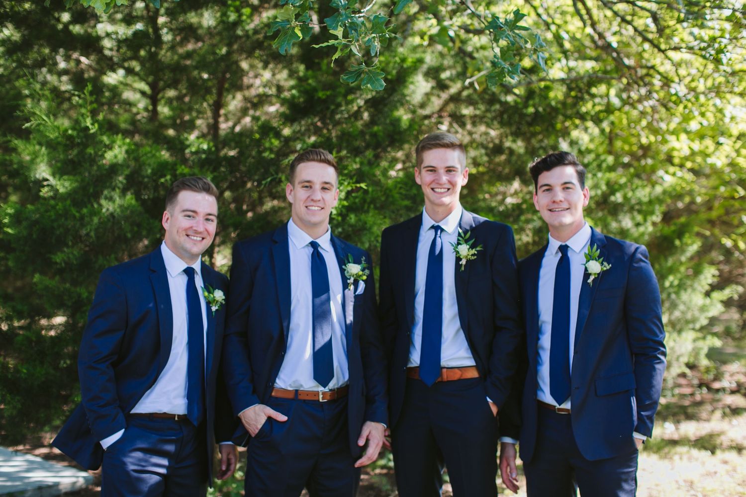 Four groomsmen in matching navy blue suits and pale blue ties pose together outdoors on a sunny day.