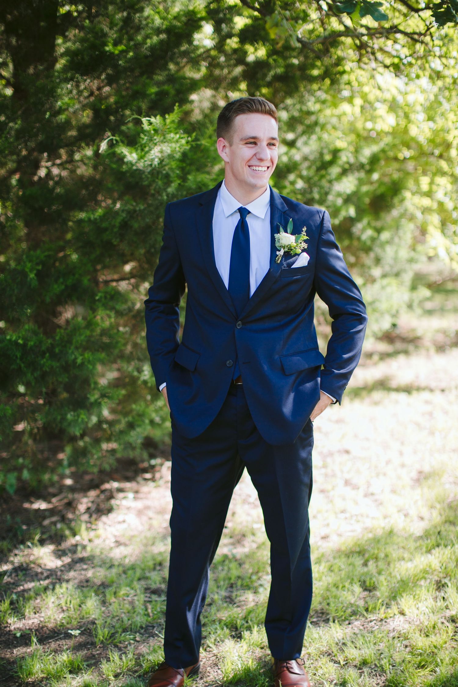 Groomsman in fitted navy suit poses with hands in pockets against natural foliage backdrop.