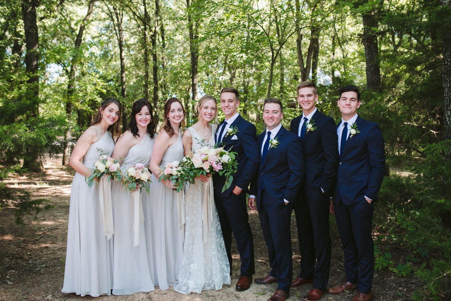 Wedding party in navy suits and light grey dresses poses together in wooded setting.
