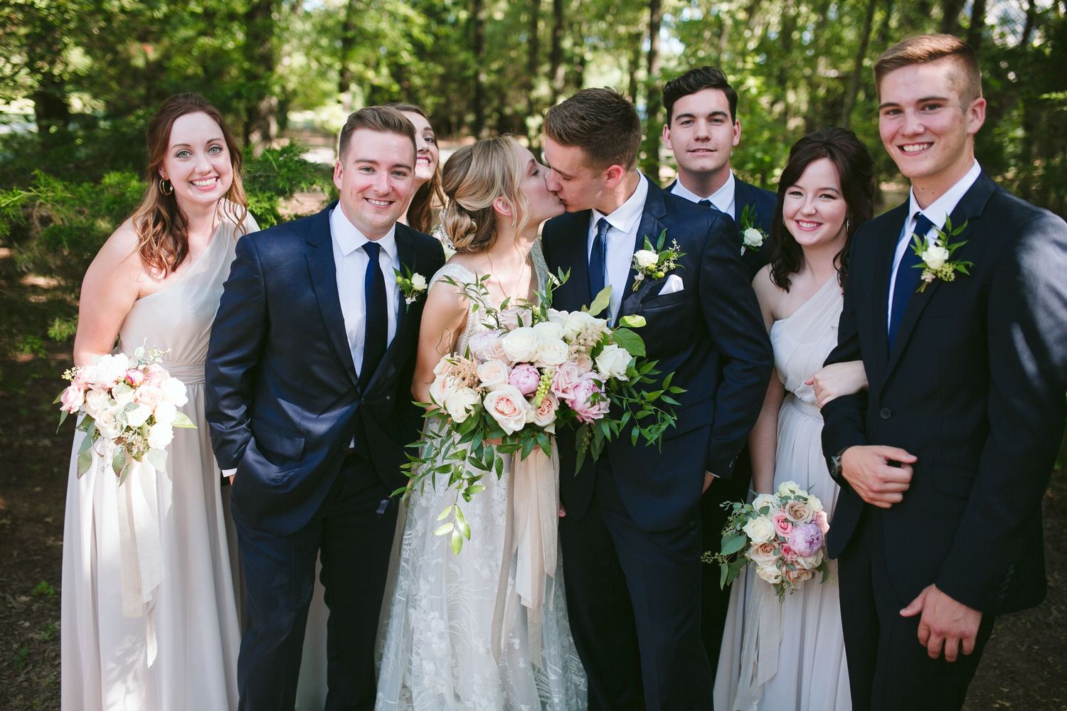 Wedding party in navy suits and blush dresses pose together outdoors with bouquets in a natural forest setting.