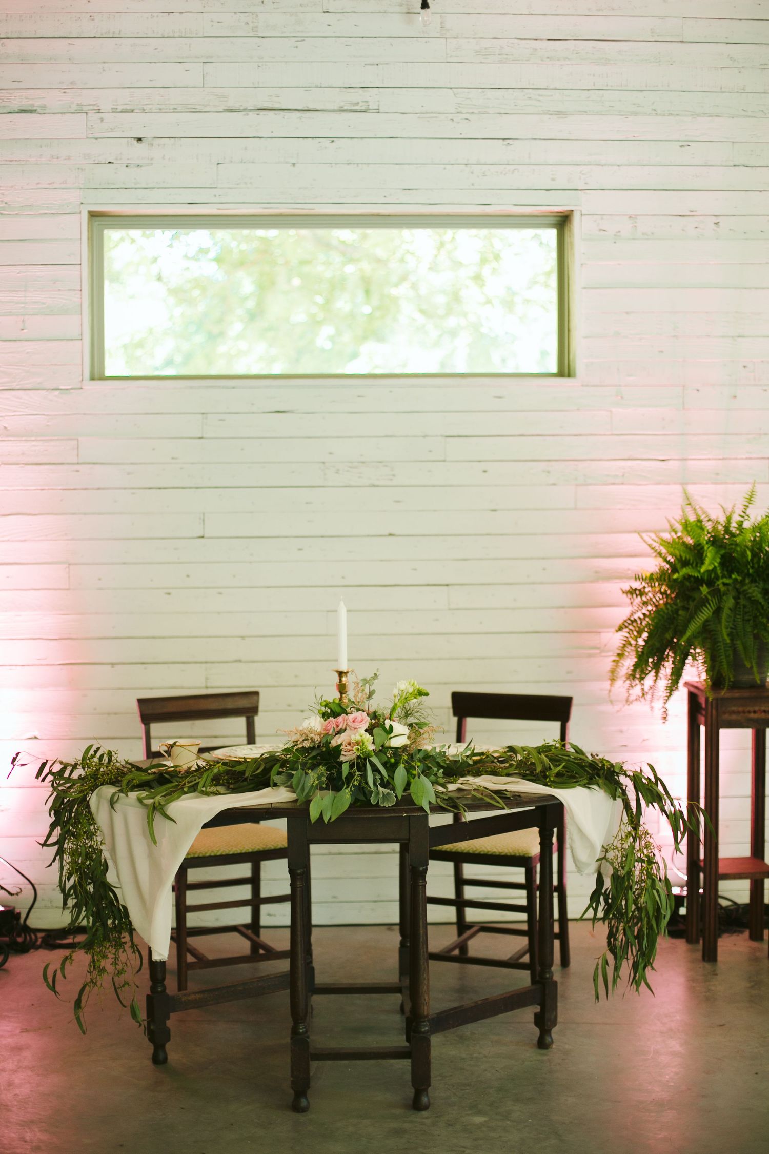 Rustic reception table setup with greenery garland and romantic lighting in modern venue.