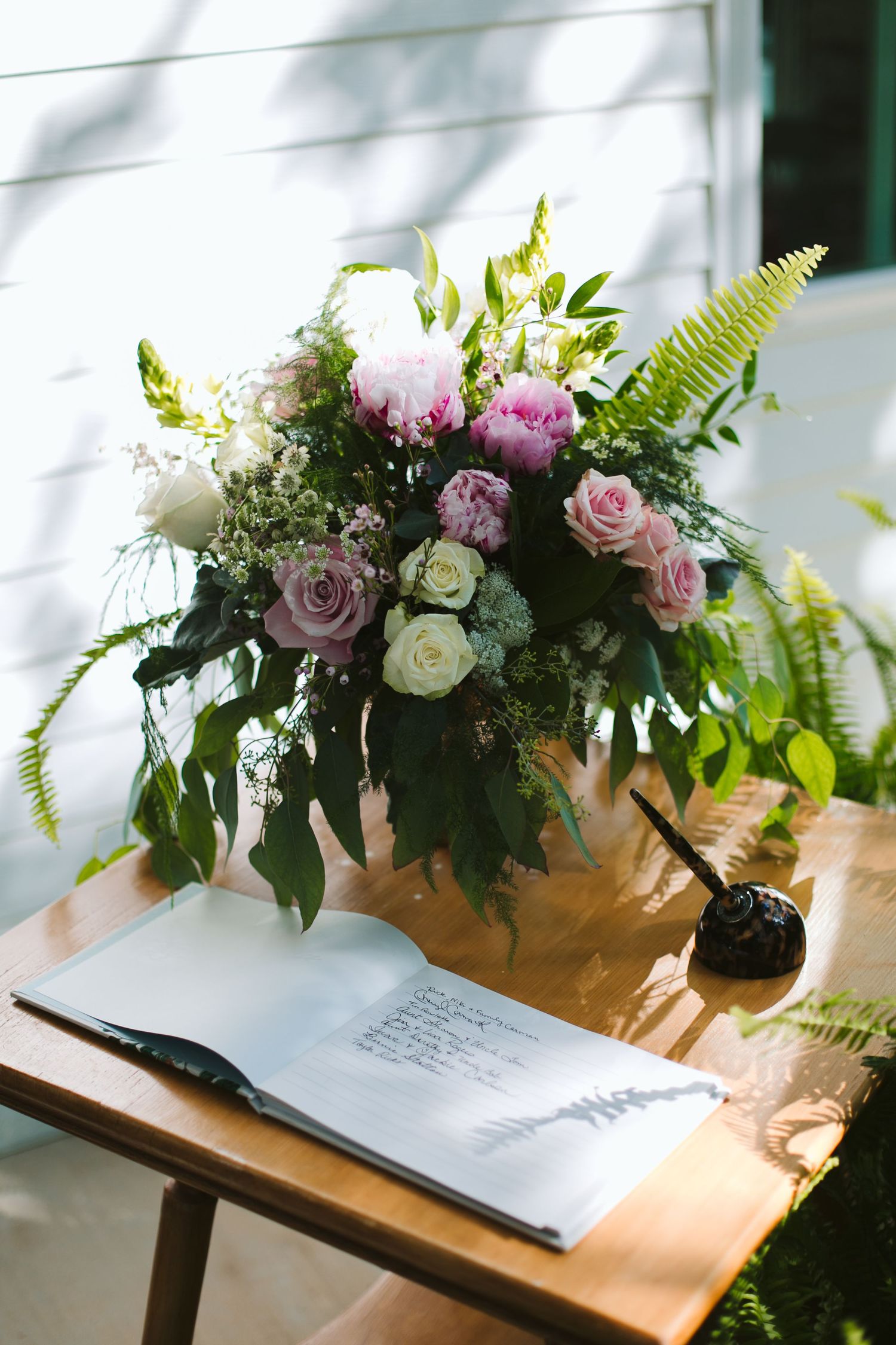 A stunning floral arrangement with pink roses and ferns sits beside an open book on a wooden table.