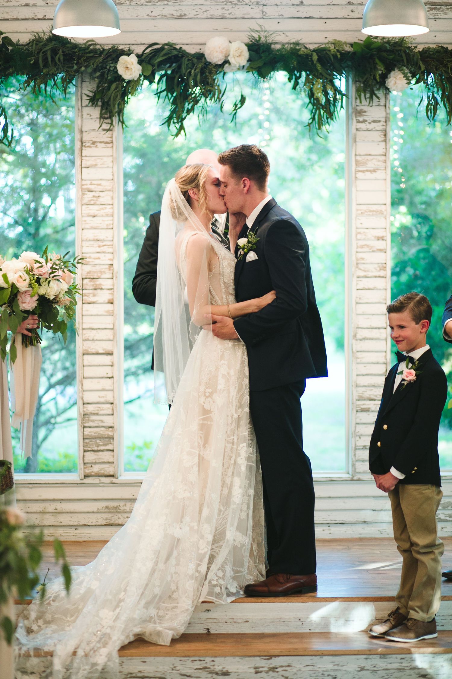 Newlyweds share their first kiss under a decorated arch while a young ring bearer watches.