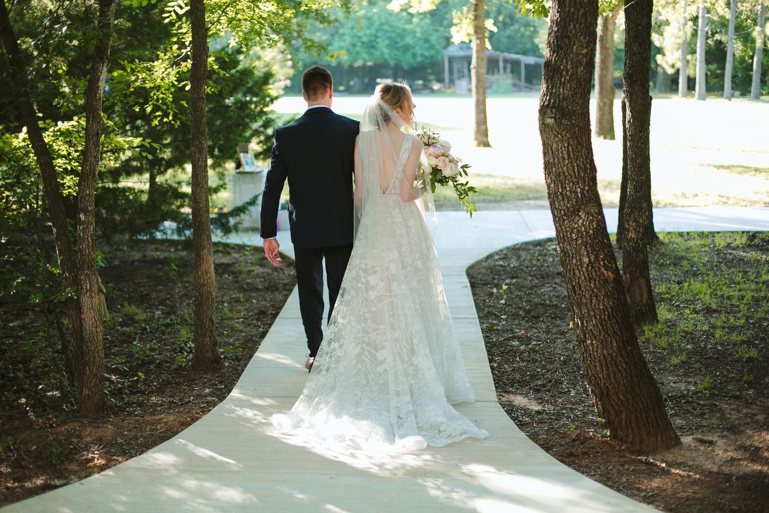 Bride and groom walking together on a winding path surrounded by trees in golden sunlight.