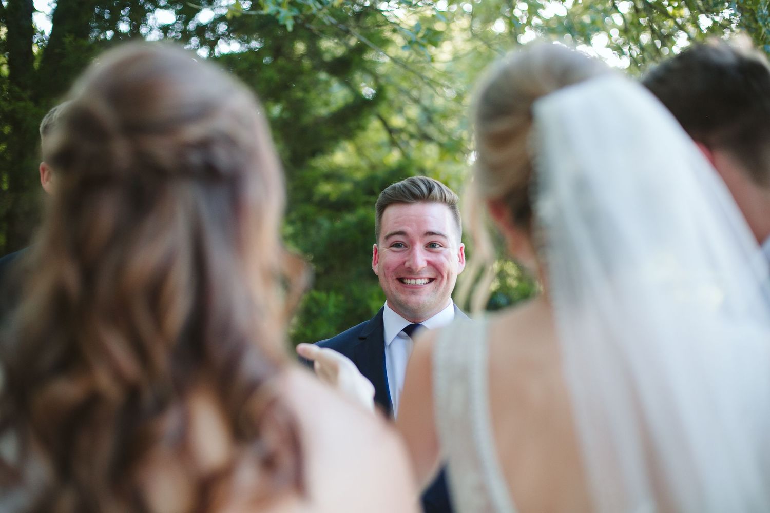 Bridesmaids in light colored dresses carry bouquets during an outdoor wedding ceremony.