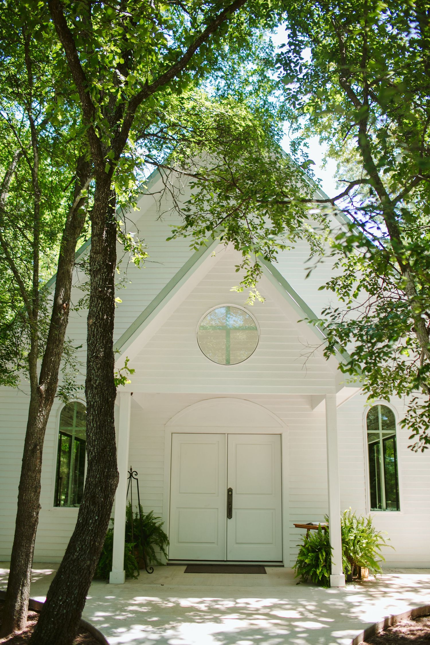 Modern white chapel with round window and double doors framed by tall green trees.