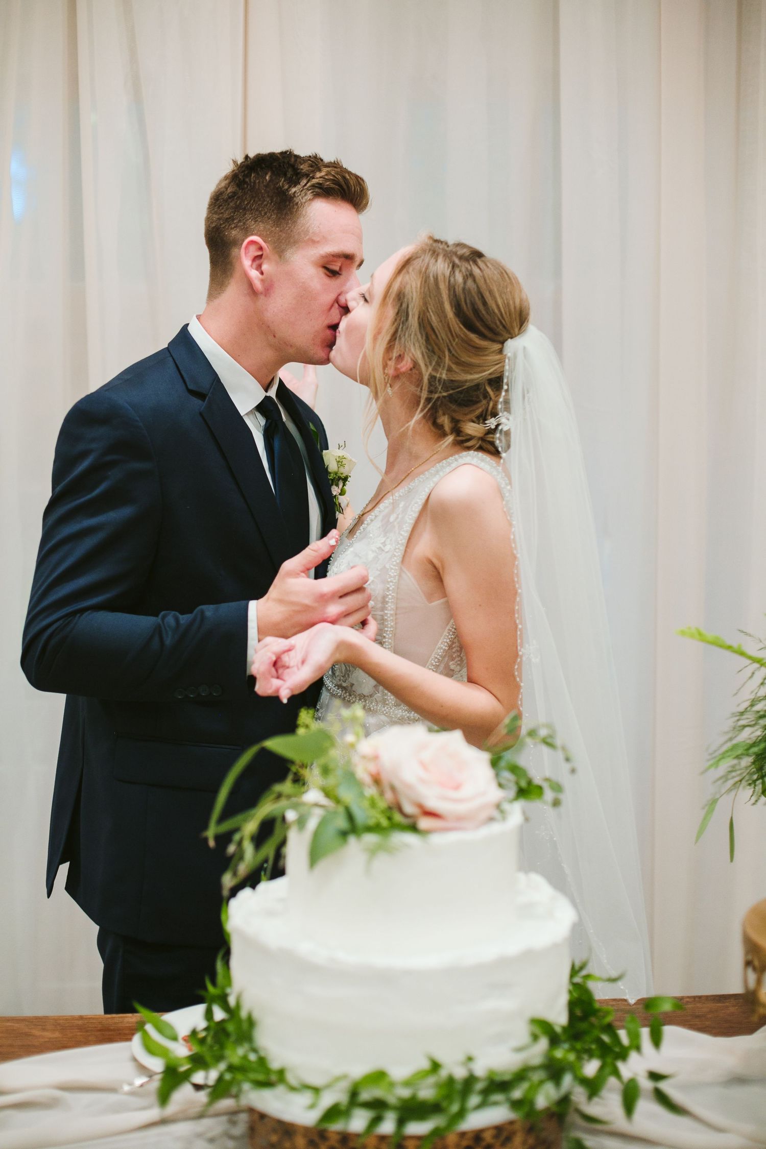 Romantic moment between newlyweds next to their white wedding cake decorated with greenery.