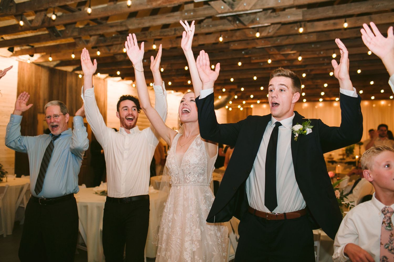 Wedding guests dance and celebrate with raised arms under rustic wooden beams and string lights at a reception.