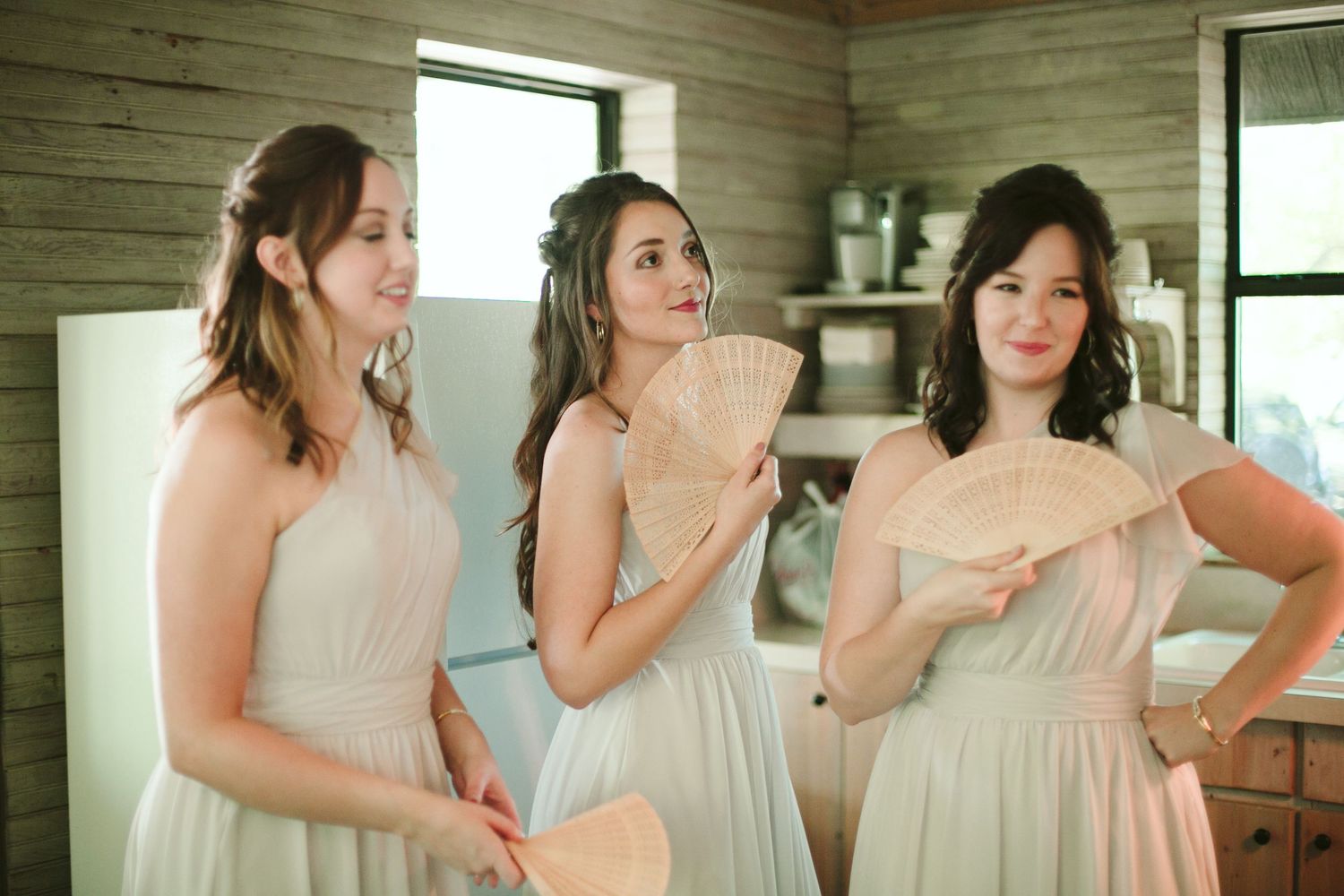Three bridesmaids in light colored dresses share a moment in rustic wooden room.
