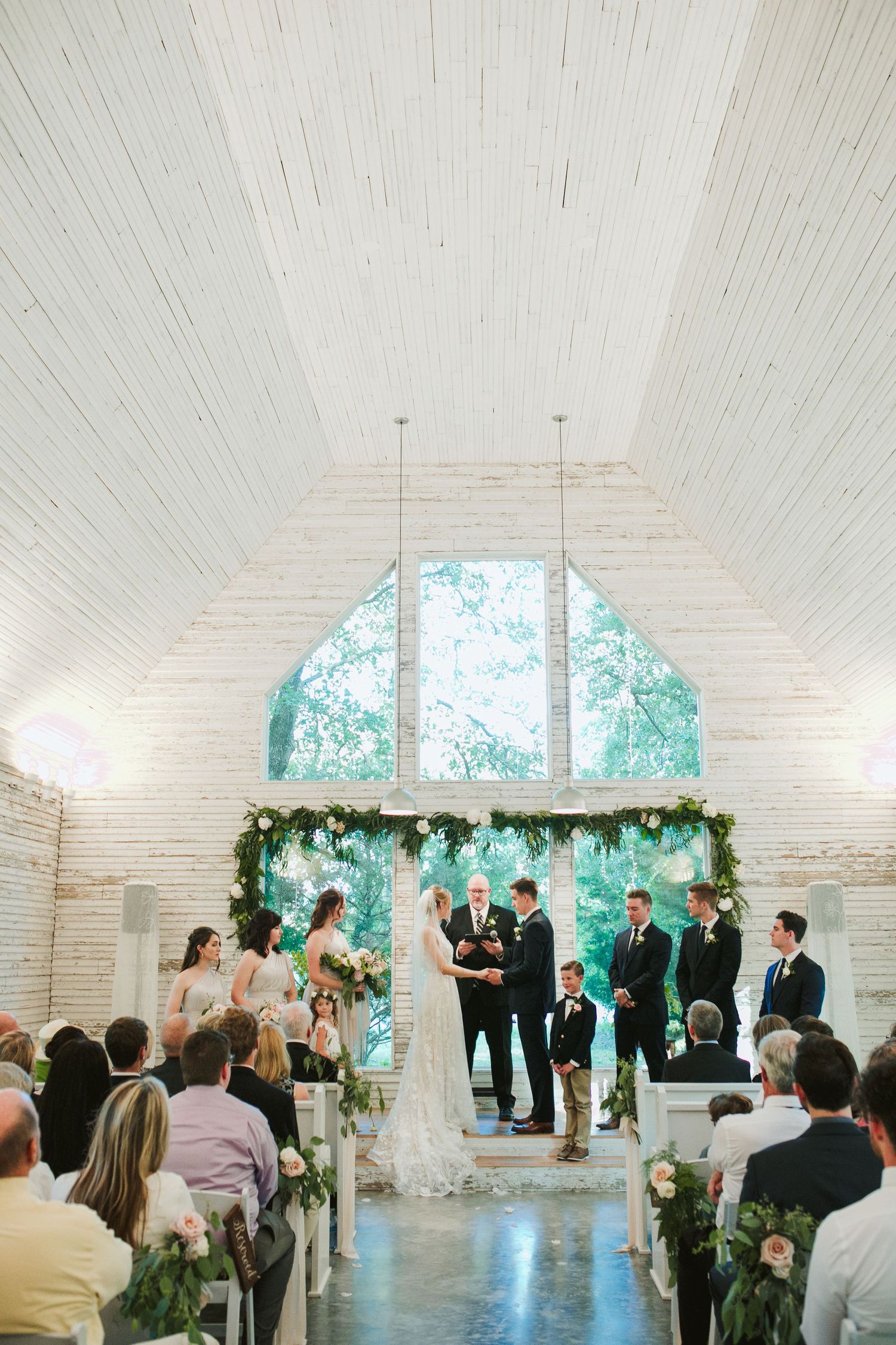 Wedding ceremony in a bright chapel with vaulted ceilings and greenery arch at the altar.