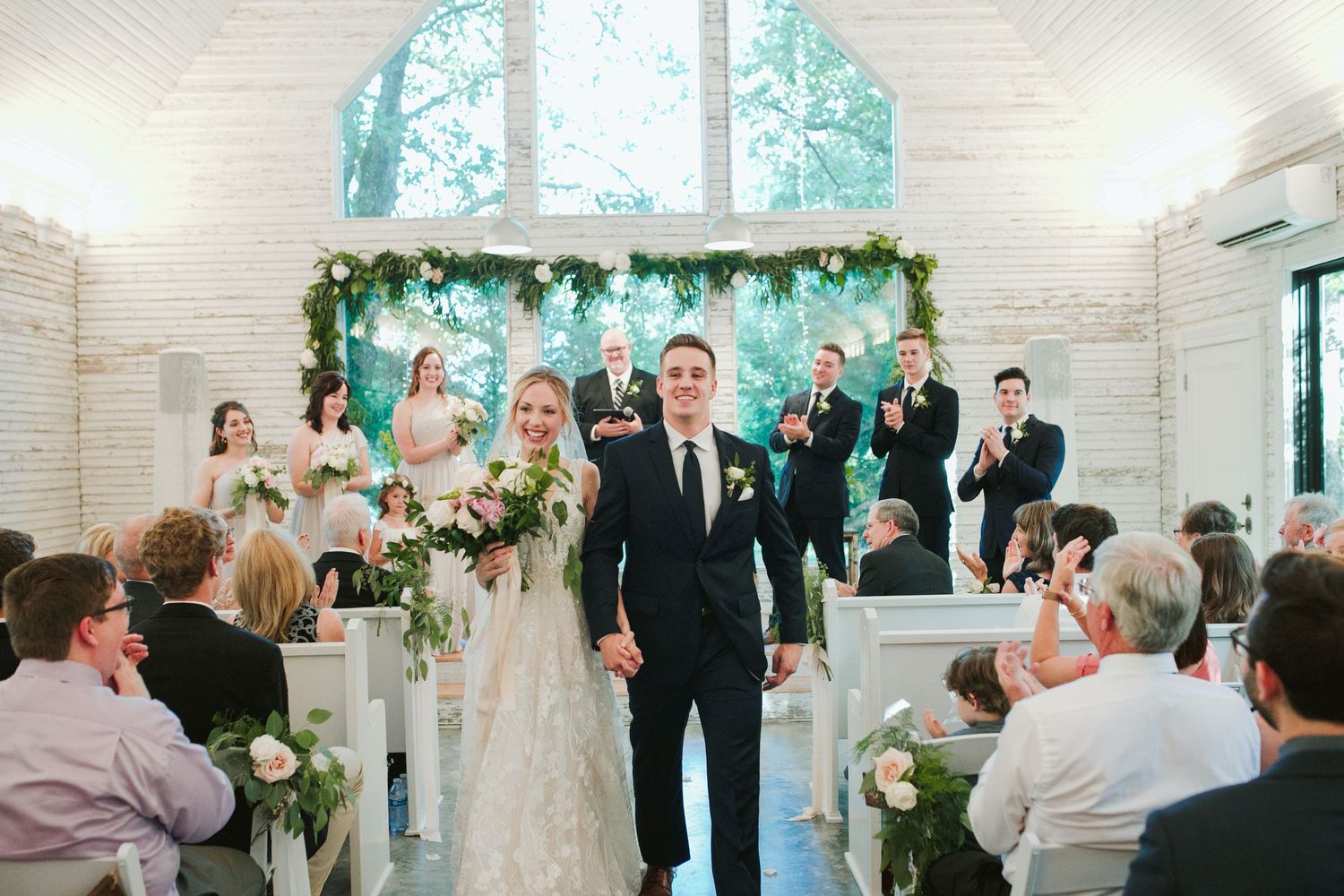 Couple walks down church aisle after wedding ceremony with floral arch and decorative greenery.