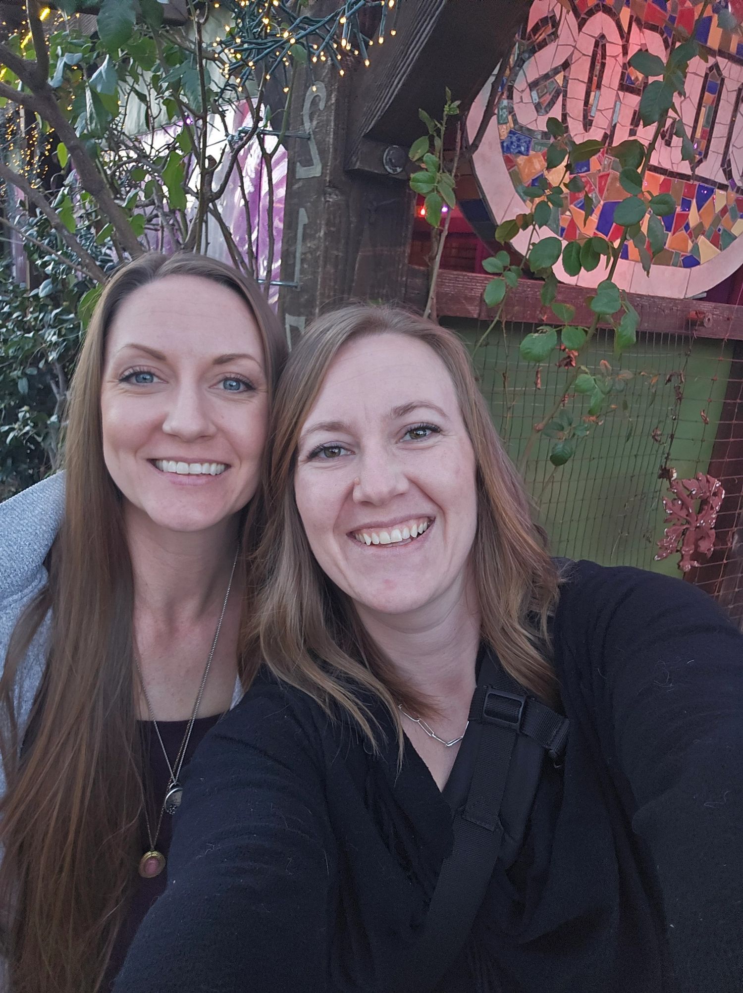 Two friends smile together in a selfie with colorful graffiti art and greenery in the background.