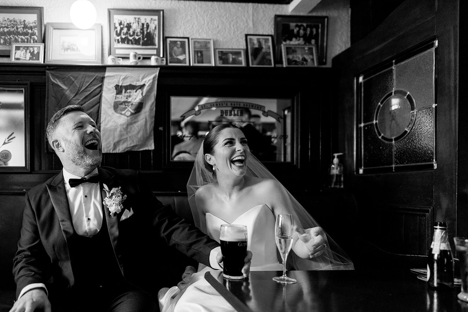 A bride and groom laugh together at a pub while sharing drinks, with framed photos decorating the wall behind them.