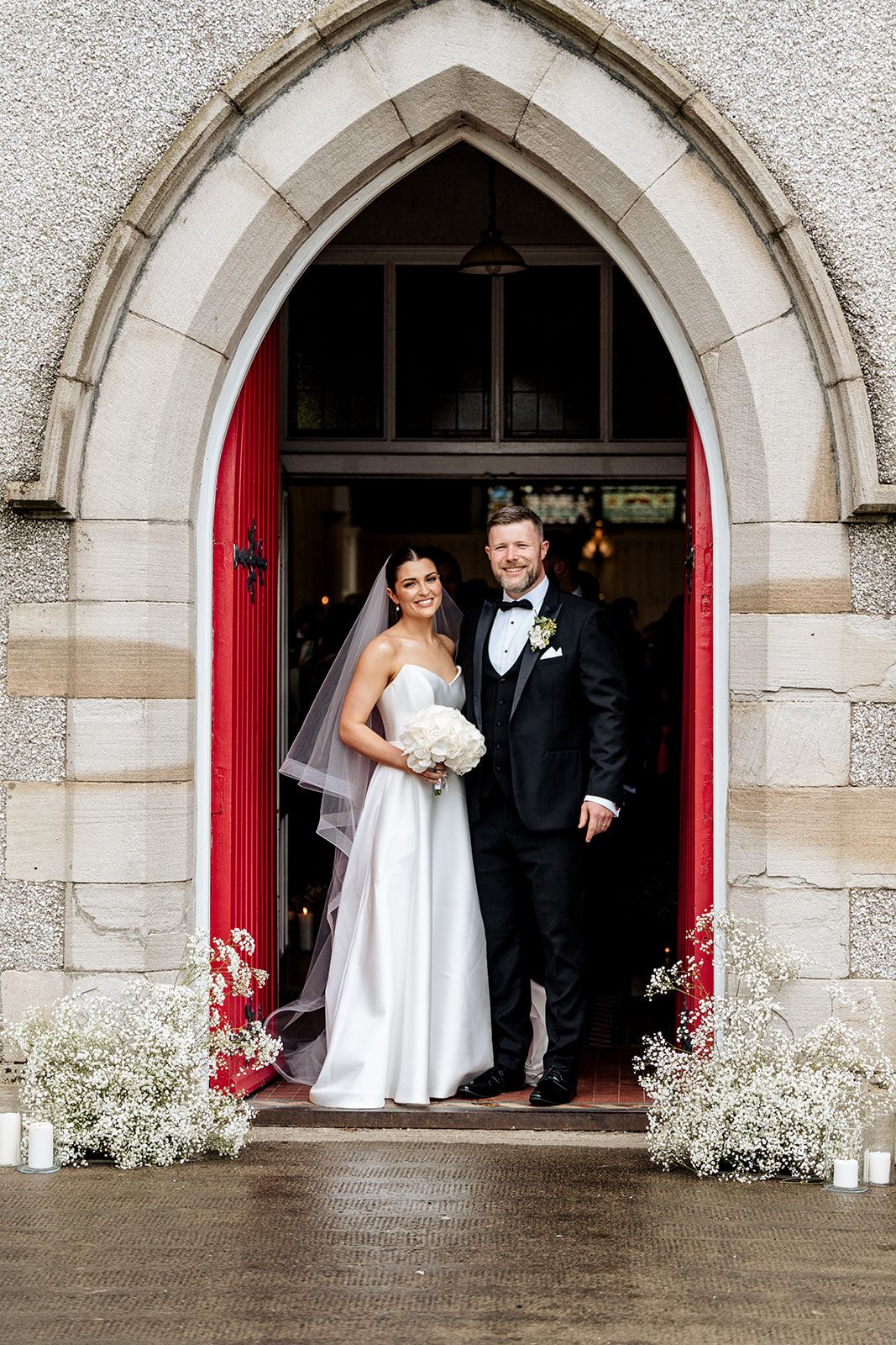 A couple stands in a stone arch church doorway with red doors, white flowers and wedding decorations.