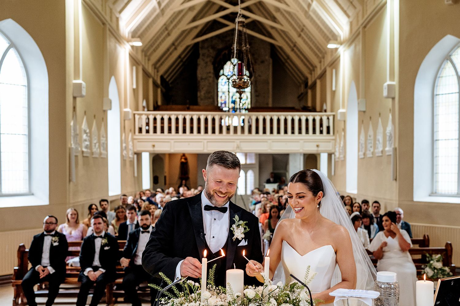Couple and guests celebrating during a wedding ceremony in an elegant white church with arched ceilings and windows.