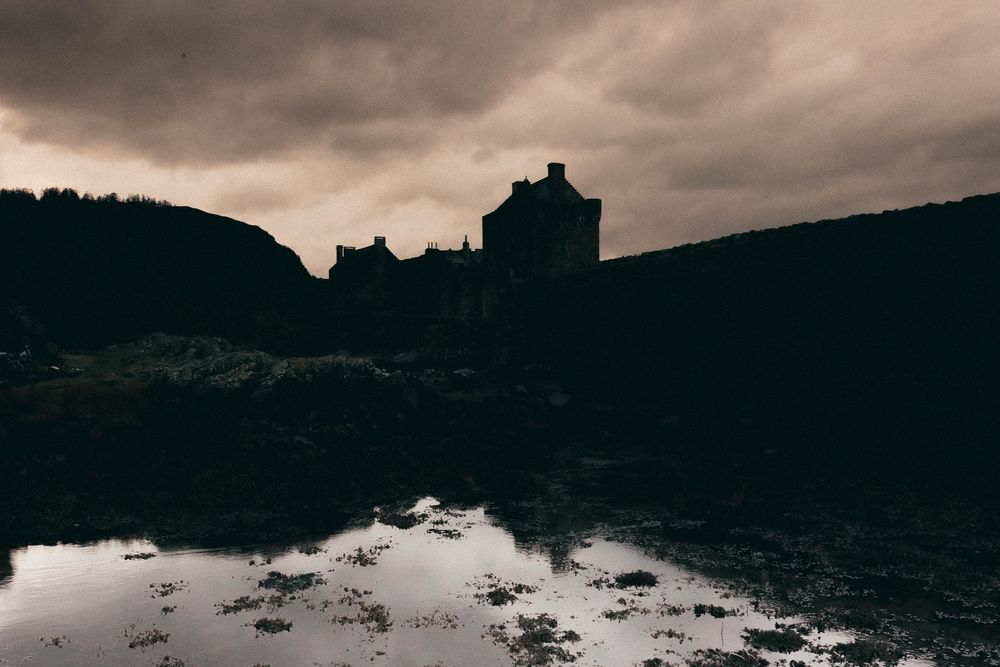 Silhouette of a castle-like structure against moody dark clouds with reflection in water below.