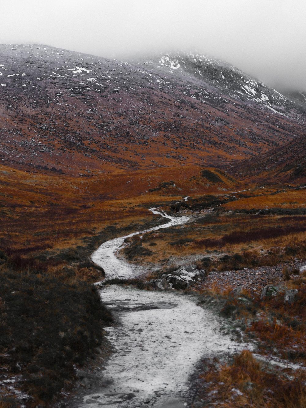 A winding river flows through autumn-colored mountains in Scotland with snow patches and misty peaks in the background.
