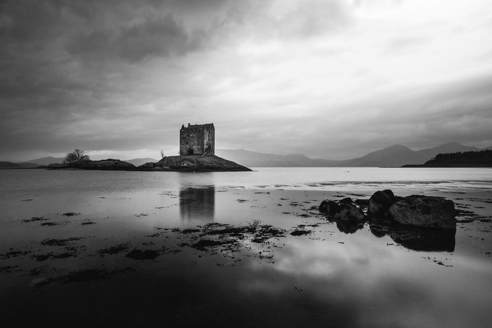 Black and white photograph of Castle Stalker silhouetted against dramatic clouds with perfect water reflection in Scotland.