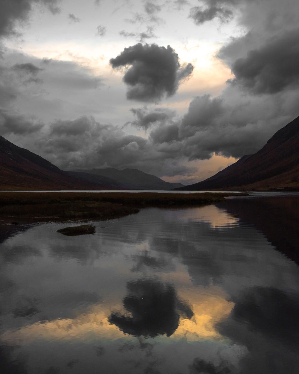 Dramatic stormy clouds reflect in still mountain lake at sunset with golden light breaking through dark sky.