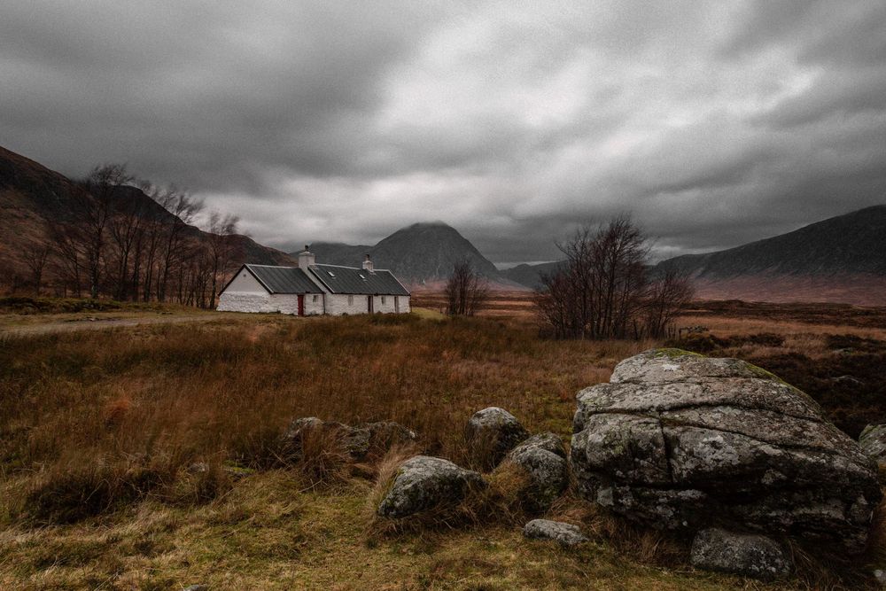 White farmhouse nestled in the Scottish Highlands under moody storm clouds with rugged rocks in foreground.