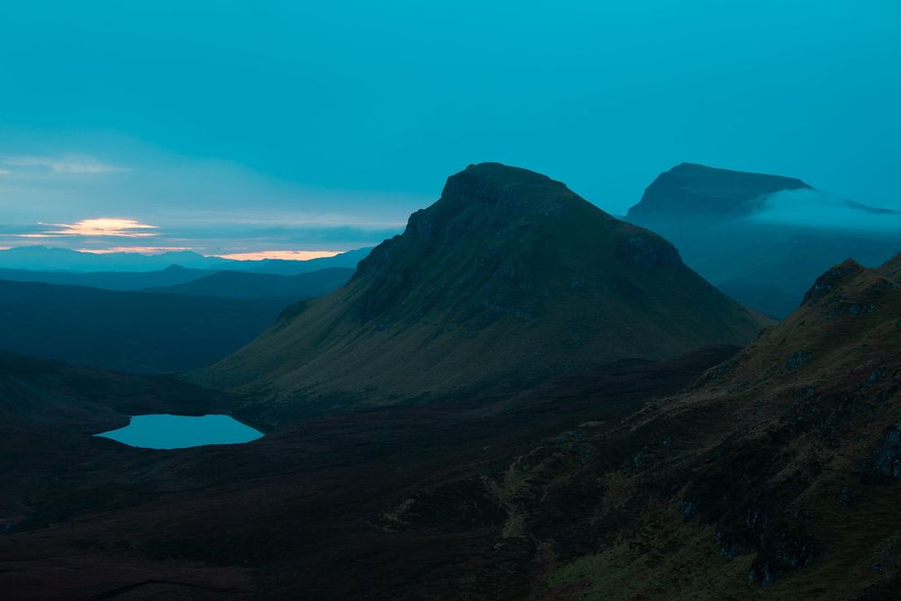 Silhouetted mountains against a vibrant turquoise sky during sunset with a calm lake in the distance.
