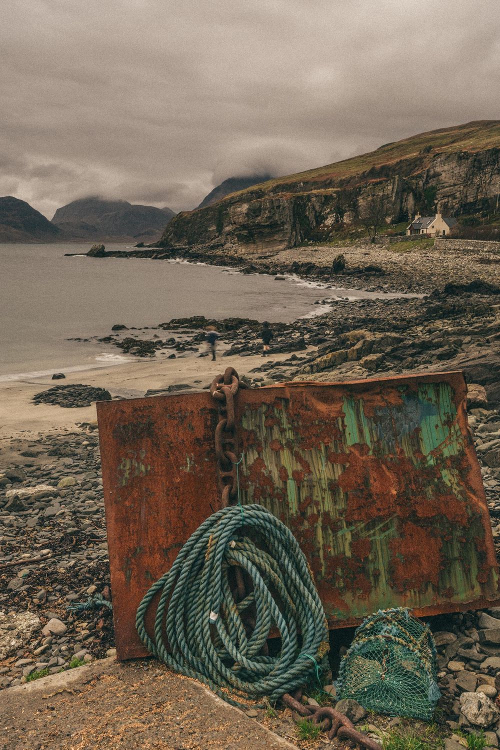 Rusty metal containers and coiled green rope on a rocky coastal shore with dramatic cliffs in the background.