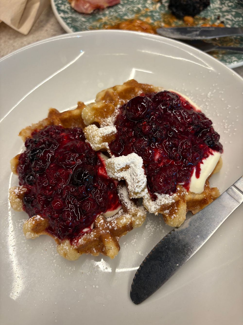 A Belgian waffle topped with dark red berry compote and powdered sugar on a white plate.