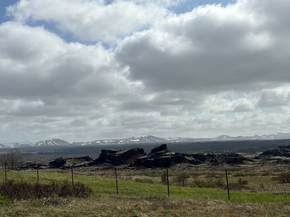 Rolling green hills stretch toward snow-capped mountains under dramatic cloudy skies in a scenic rural landscape.