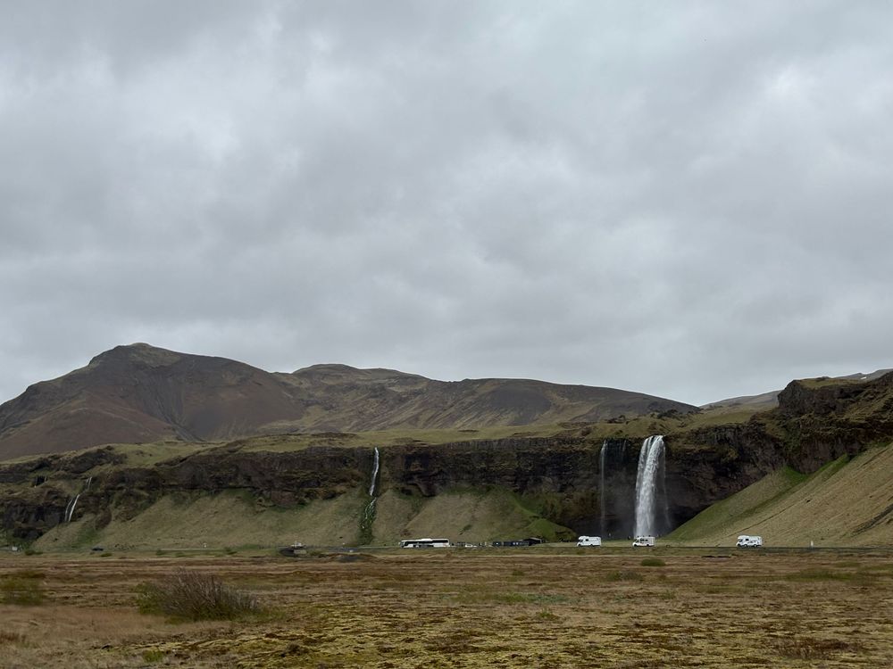 A misty waterfall cascades down green cliffs in the rugged Icelandic landscape under overcast skies.