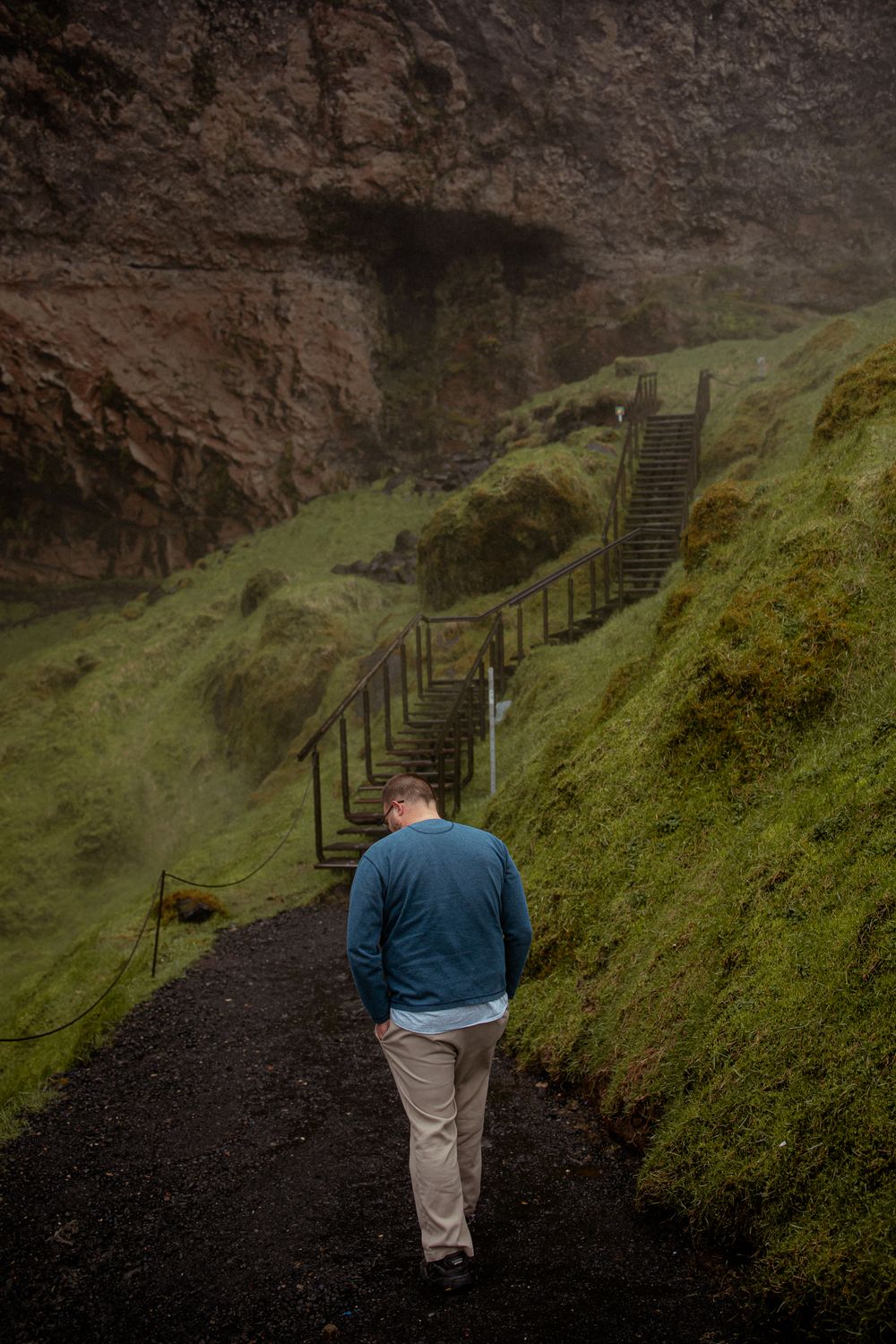 A lone figure walks along a dark, mossy path toward wooden stairs leading up a steep green hillside in Iceland.