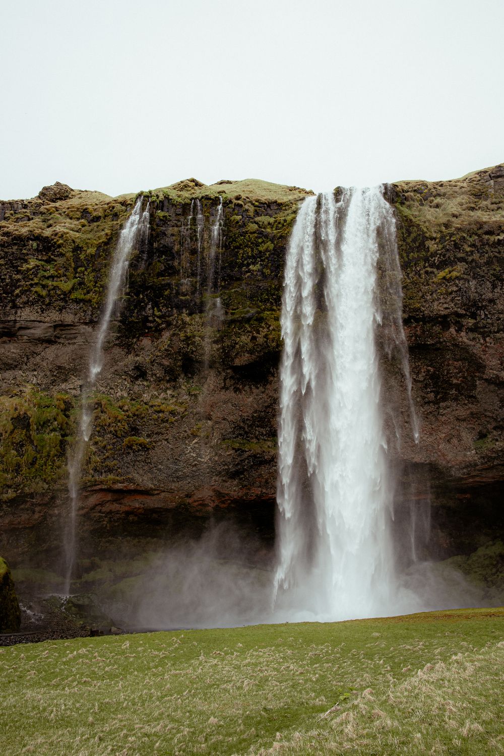 Majestic waterfall cascades down a moss-covered cliff face onto green grass in Iceland's rugged landscape.