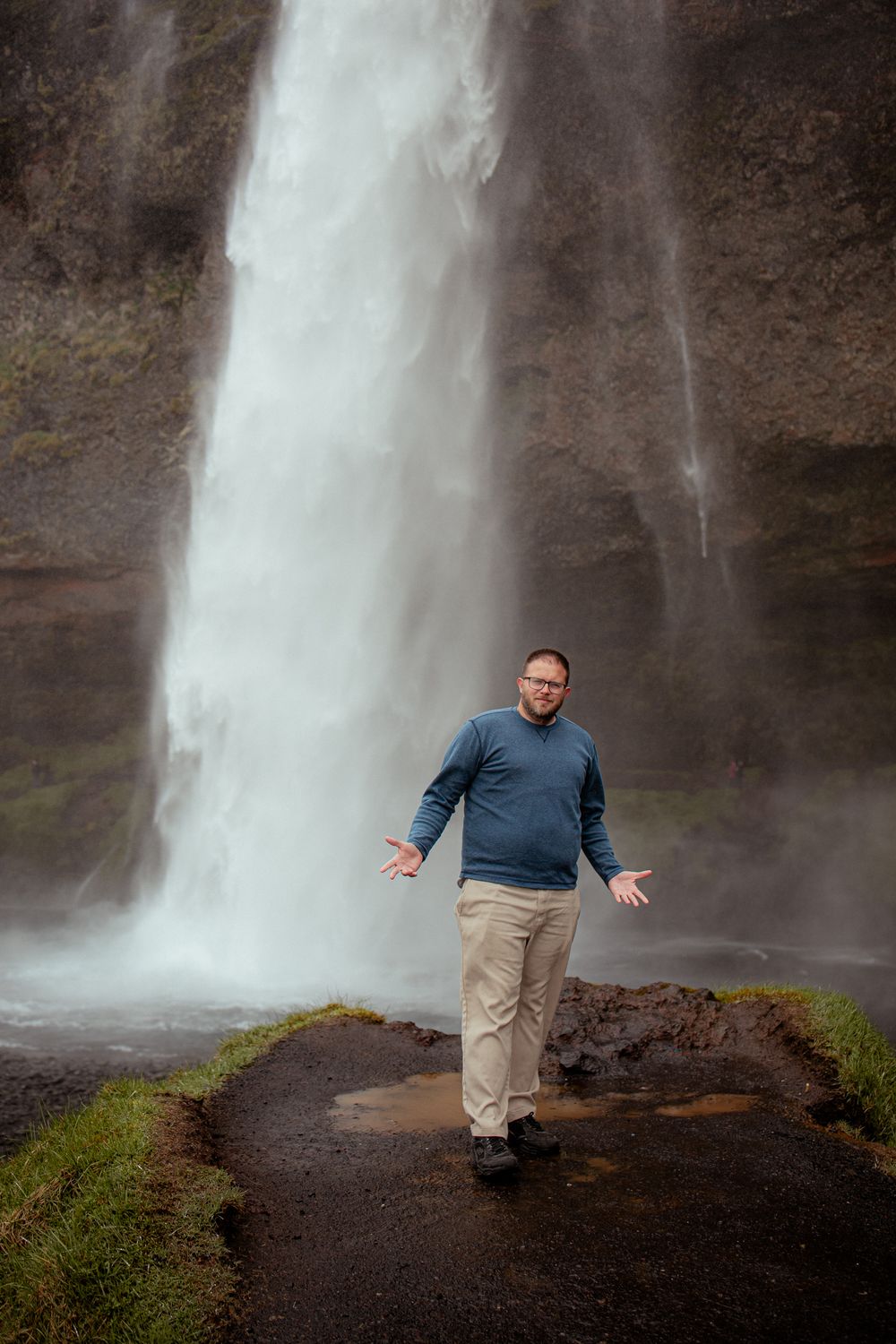 A person gestures with arms outstretched near the base of a towering waterfall on a misty hiking trail.