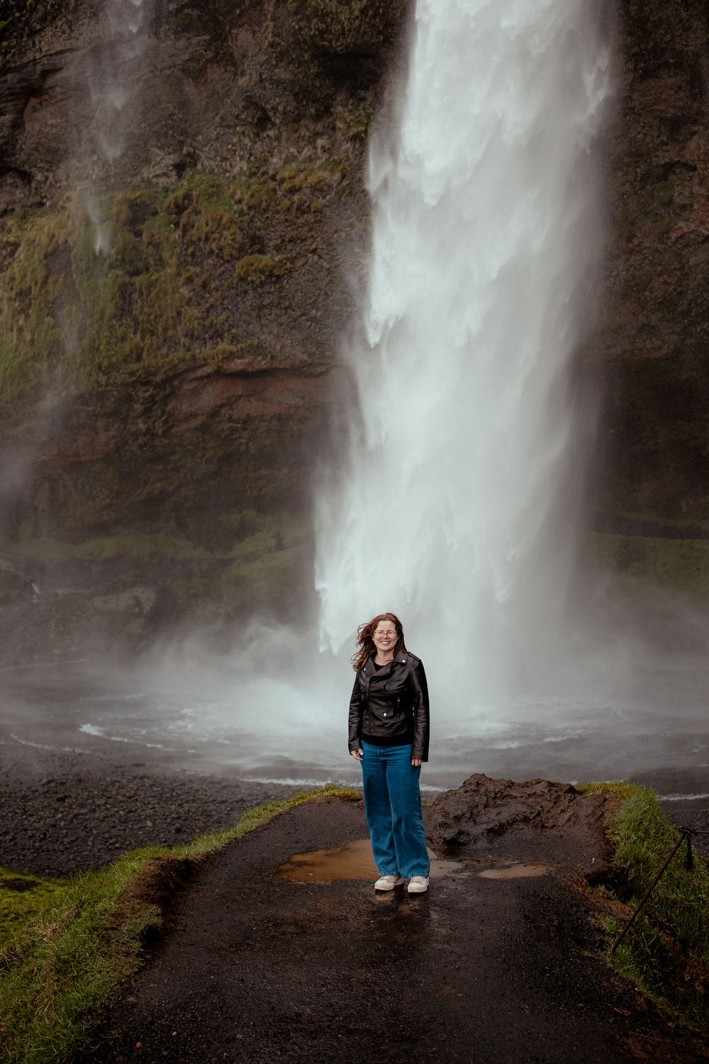 A person stands on a dark path beneath a majestic waterfall cascading down moss-covered cliffs in misty conditions.