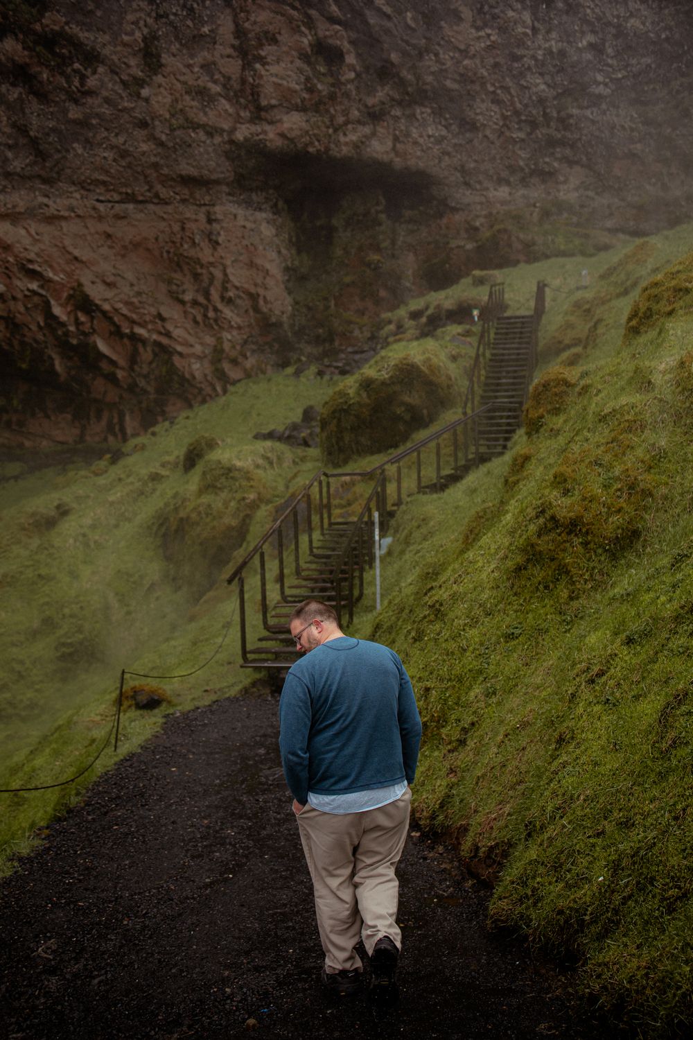 A hiker in a blue sweater walks along a misty volcanic trail with wooden stairs ascending the mossy hillside in Iceland.