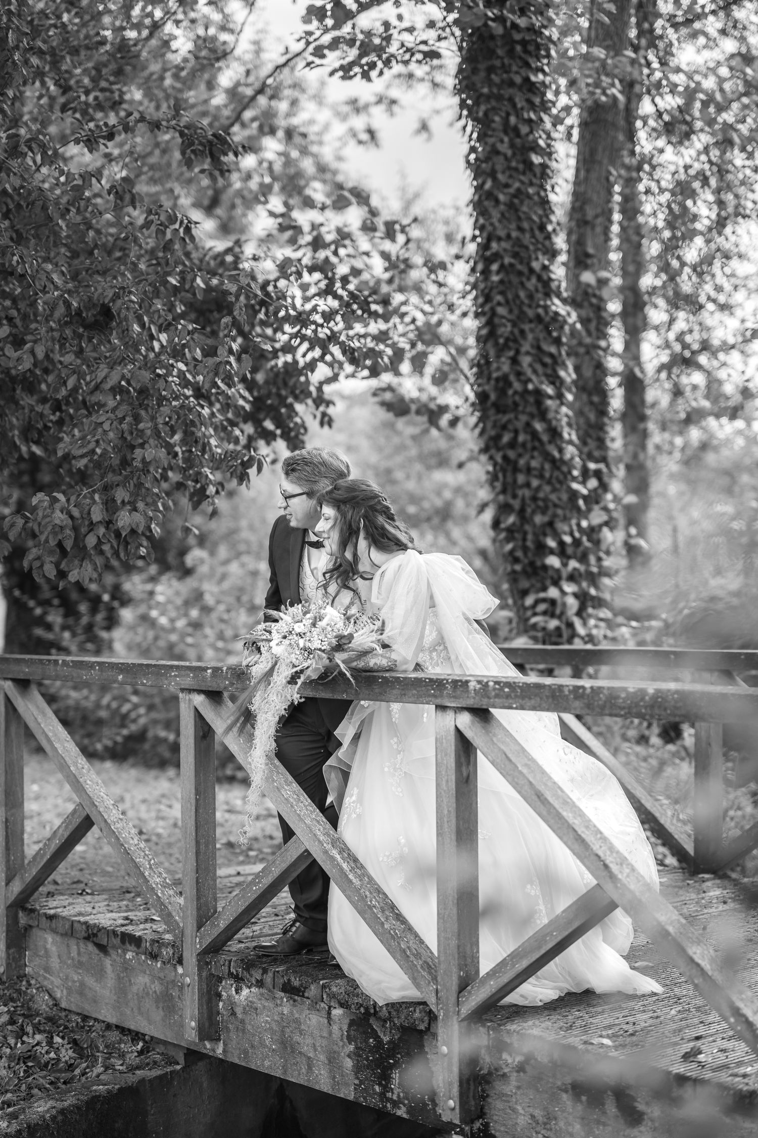 Romantic black and white wedding photo of couple sharing intimate moment on rustic wooden bridge surrounded by lush foliage.