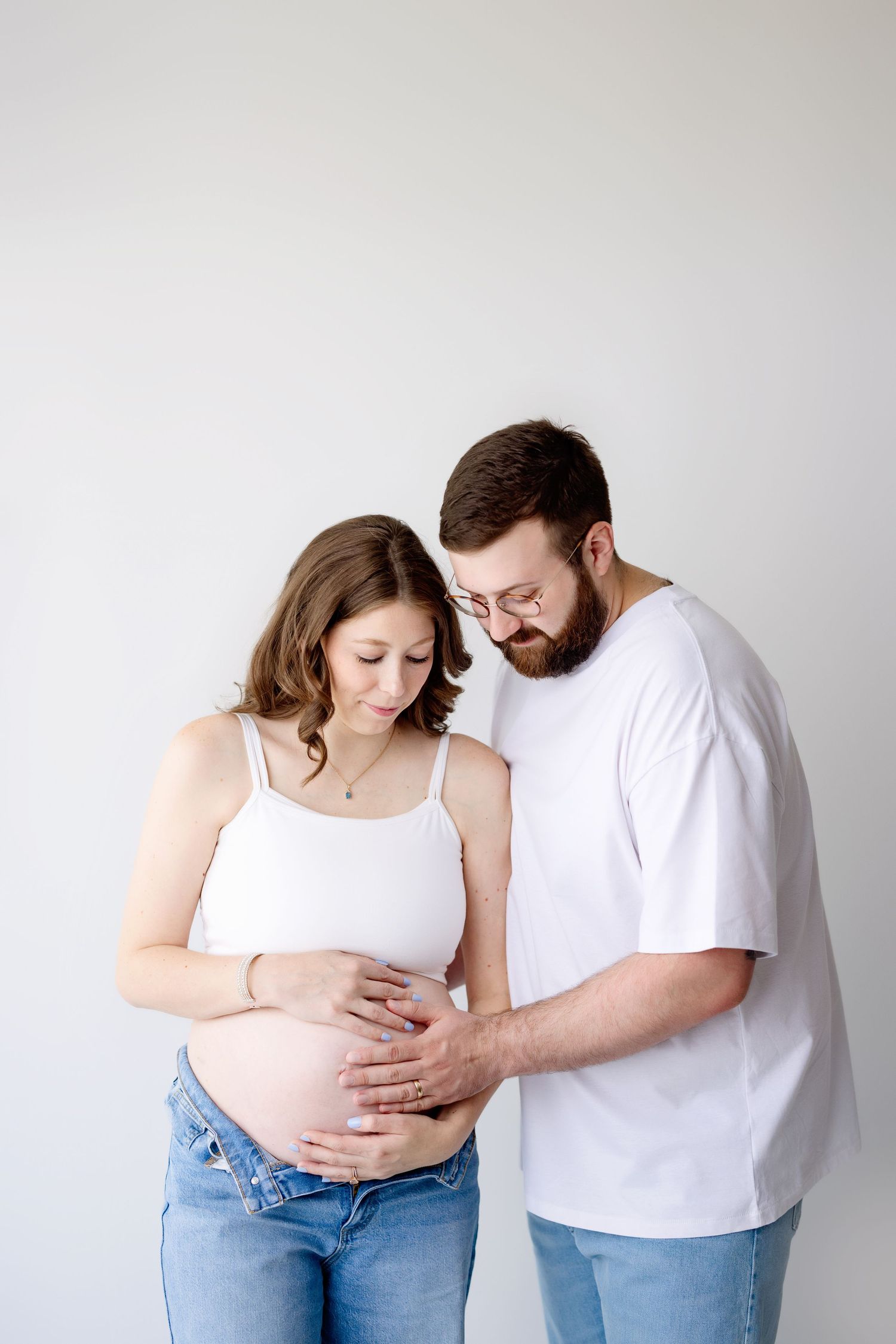 Expecting parents in white tops and jeans share tender moment embracing baby bump against neutral wall background.
