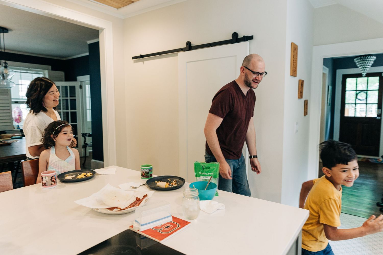 People gather around a bright white kitchen counter with plates and food during a casual mealtime at home.