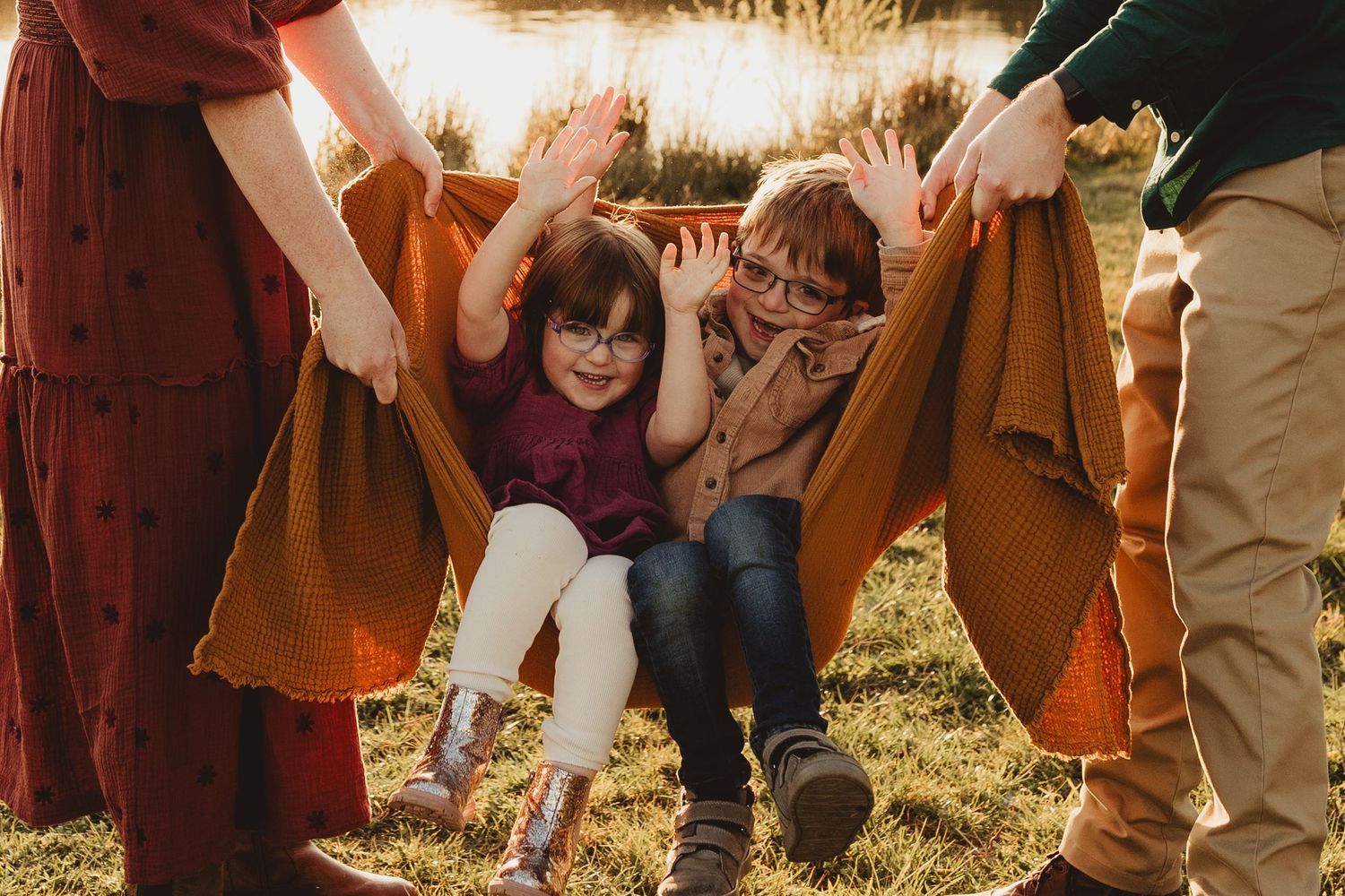 Two children smile and play under a brown blanket being held up by adults in autumn sunlit field.