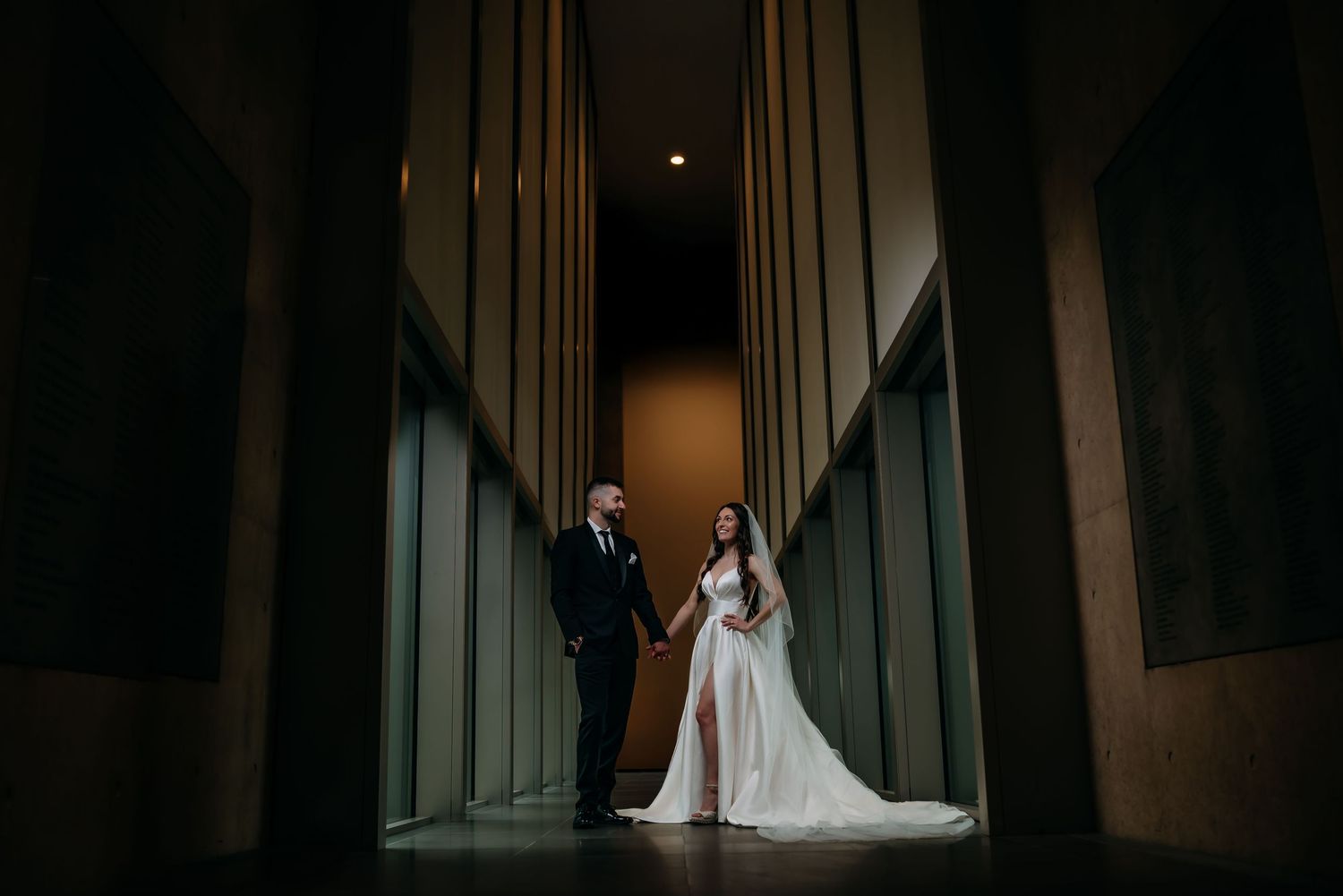 A couple in formal wedding attire stand dramatically in a long hallway with tall architectural columns in the Grand Rapids Art Museum