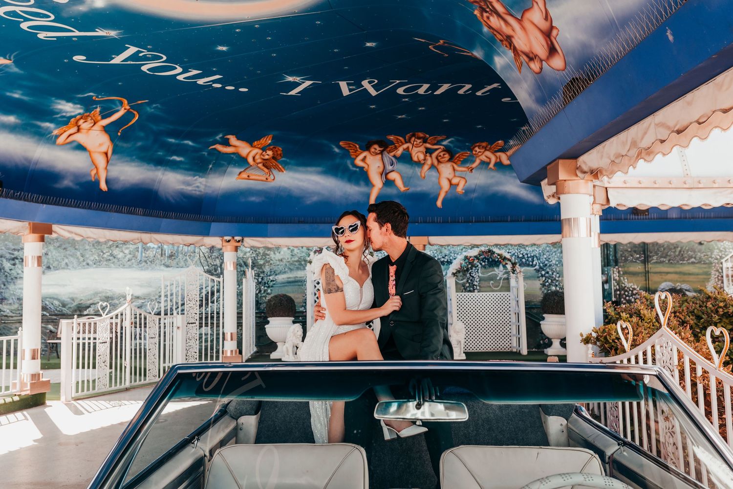 A romantic couple sits atop a vintage car under a whimsical blue ceiling decorated with cherub figures.