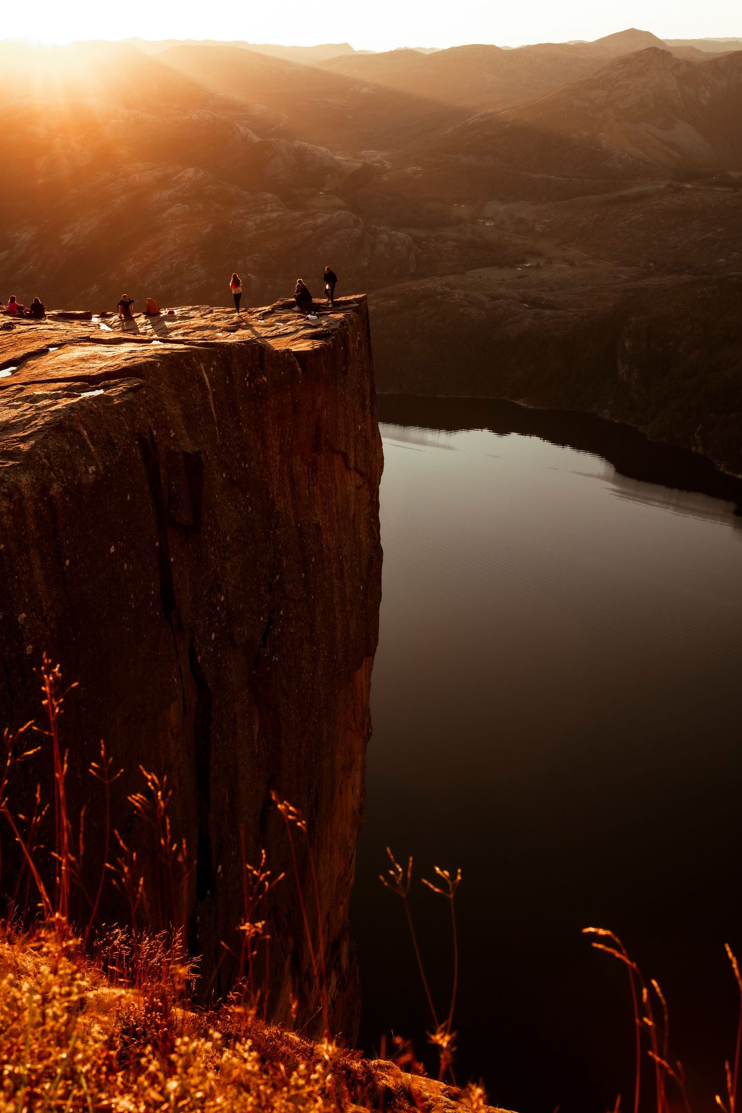 Silhouetted figures stand on dramatic cliffside overlooking serene lake at sunset in mountainous landscape.